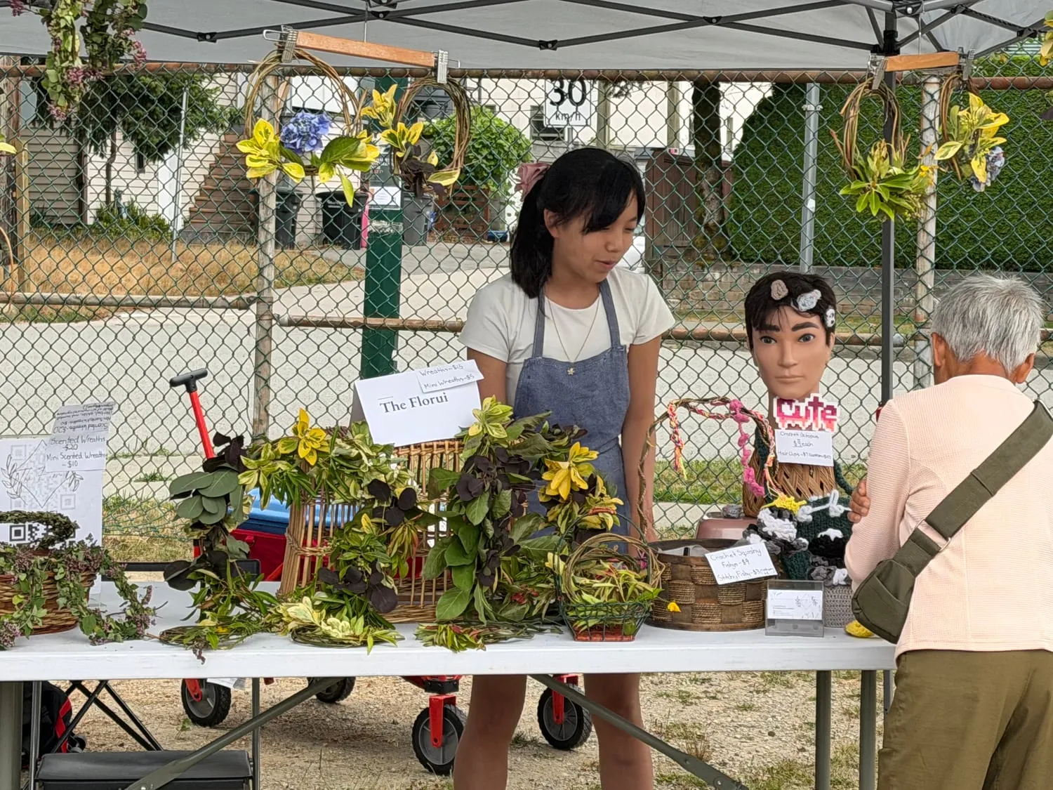A young entrepreneur at The Florui's table, arranging garden wreaths beside a hand-lettered price list.