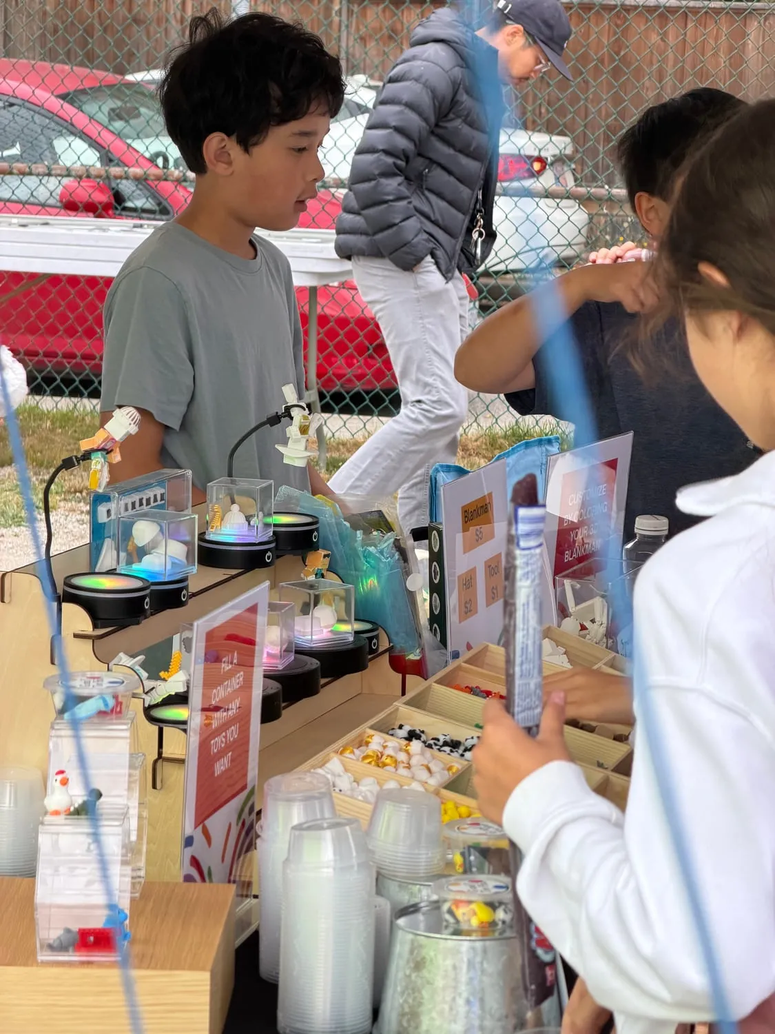 A young entrepreneur behind a table of 3D-printed toys in lit display cases, with printed signs offering to customize a figure by colouring it.