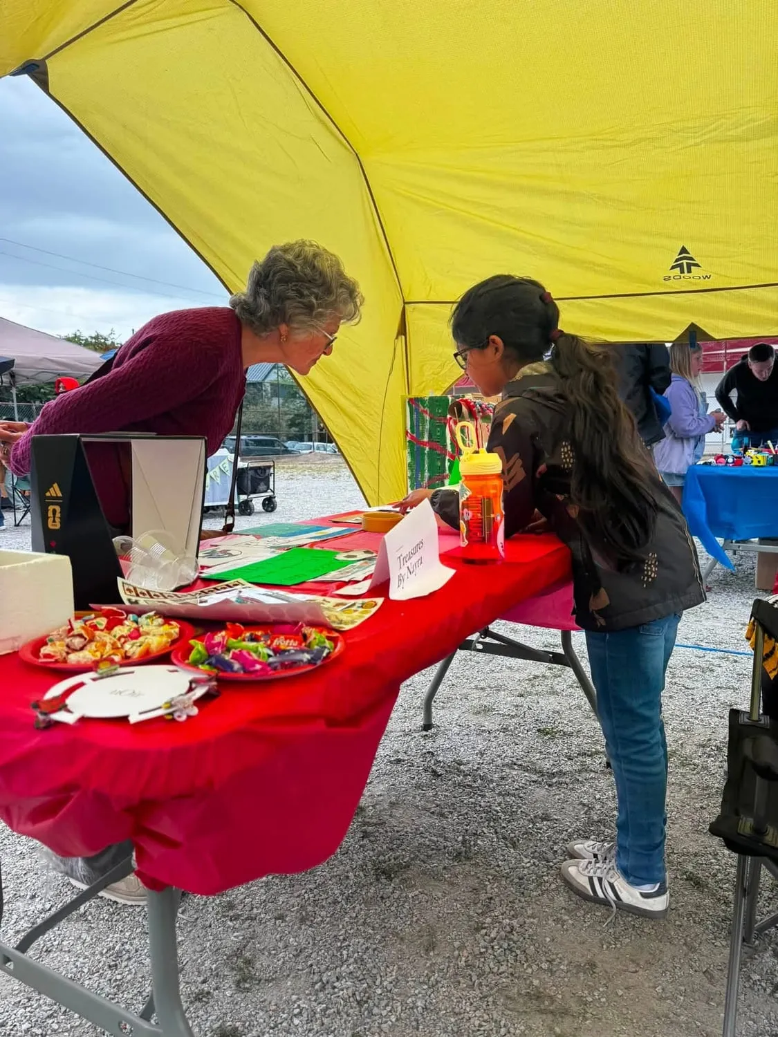 A child and an adult lean over a red market table with handmade paper goods beneath a yellow canopy.